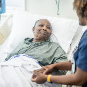 A female nurse of African decent checks in on her senior patient as she awaits to be called for surgery. She is smiling as she reassures her patient and puts her at ease before the procedure.