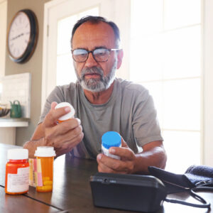 A Latino senior citizen sits at his dining room table and sorts through various prescription medications as sunlight filters in through the window behind him bathing the room with a soft glow of light. A blood pressure machine rests on the table in front of him.