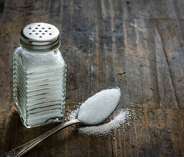 Glass salt shaker shot on rustic wooden table. A spoon with salt is beside the shaker placed directly on the table. The composition is at the left of an horizontal frame leaving useful copy space for text and/or logo at the right. Predominant colors are white and brown. High resolution 42Mp studio digital capture taken with Sony A7rII and Sony FE 90mm f2.8 macro G OSS lens